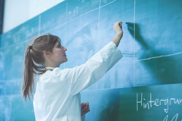 A woman wearing a white lab coat writing on a blackboard